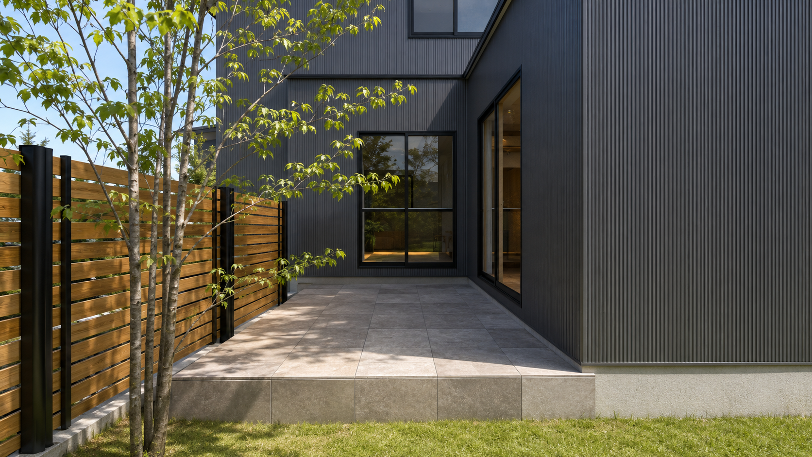 Modern black corrugated exterior house with large windows, a wooden slat fence, and a tiled patio courtyard.