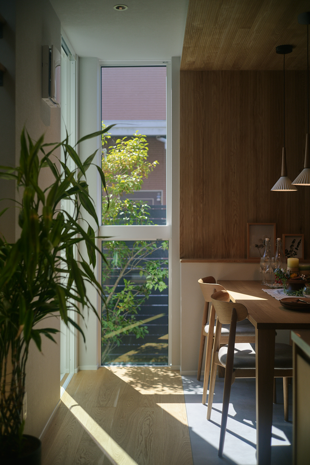 Sunlit dining area with a wooden table and matching chairs beside a large window, potted plant in the foreground, and hanging pendant lights on a wood-paneled wall.