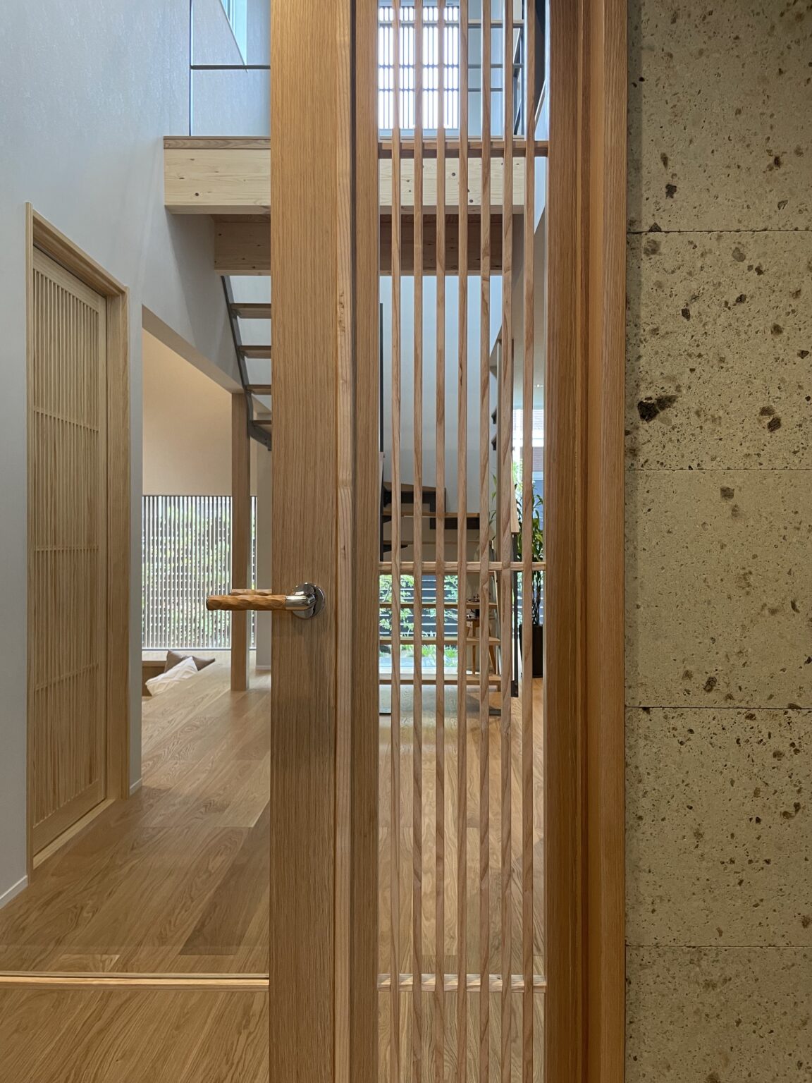 Interior view through a vertical wooden slat screen into a sunlit living area with a wooden staircase in the background.