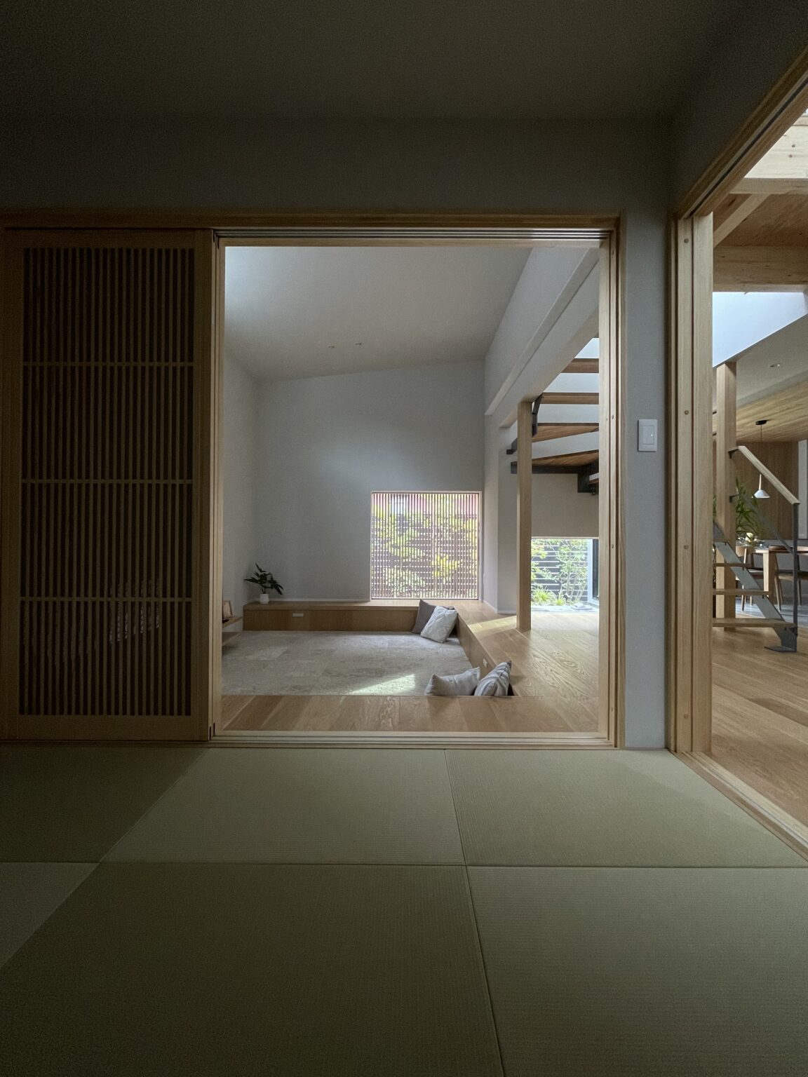 Dark foreground tatami room opens to a bright, minimalist living area with light wood beams and stairs beyond the opening.