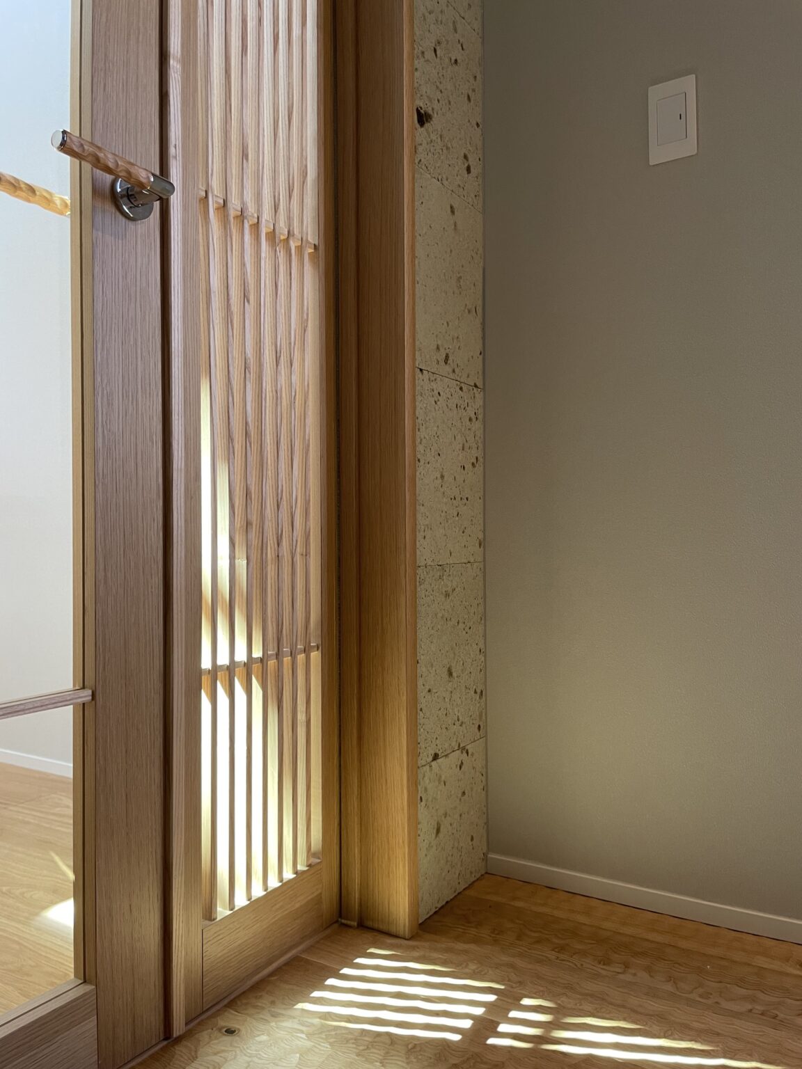 Wooden slatted room divider with light streaming through gaps, casting striped shadows on a wooden floor near a beige wall and stone column.