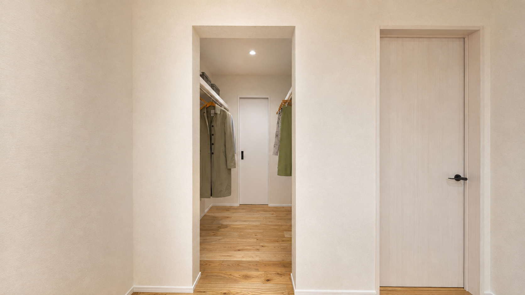 Open closet in a hallway showing hanging coats, wooden floor, and a small recessed light in the ceiling. There’s a white door at the end of the closet.